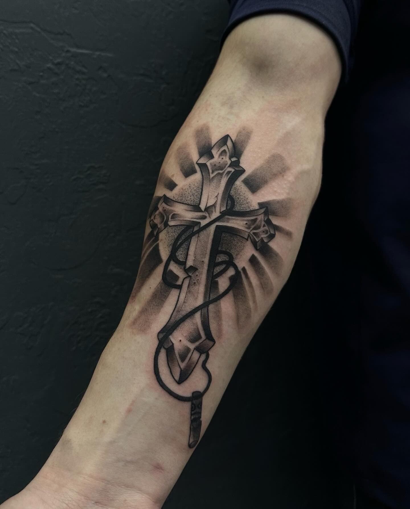 Black and grey cross tattoo with rays of light on the forearm